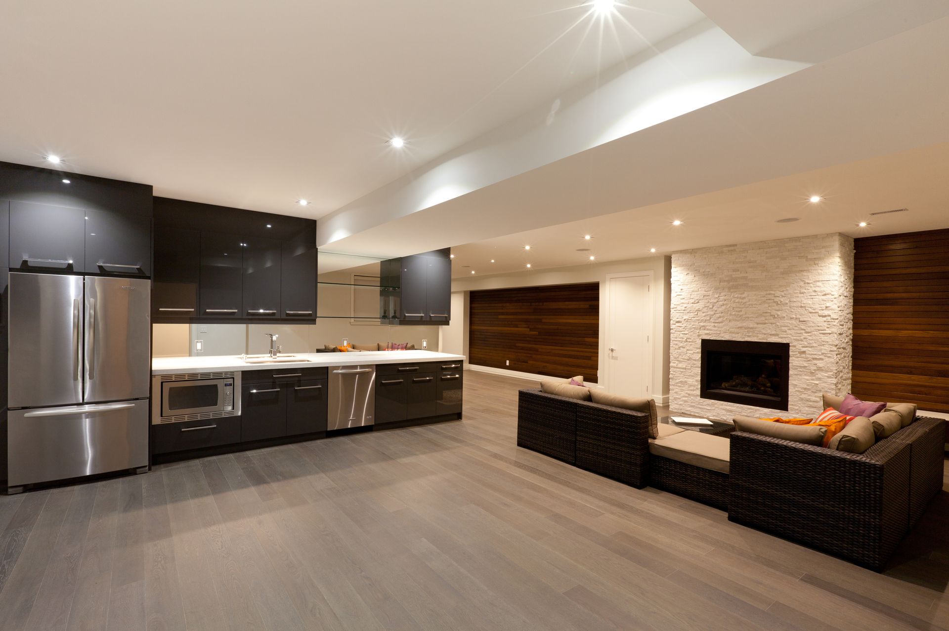 Modern basement kitchen and living area. Features a stainless steel fridge and microwave, dark cabinets, a fireplace, and wooden accent walls.