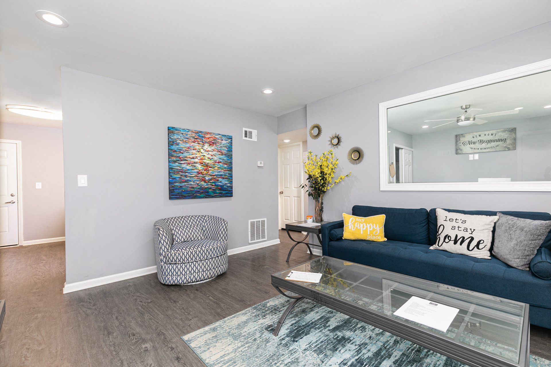 Apartments in Forest Park, GA - Gardens at Park West - Furnished Living Room with Vinyl Plank Flooring, Blue-Grey Area Rug, Navy Sofa, White and Black Armchair, Rectangular Glass Coffee Table, Soft Grey Walls, Wall Art, Framed Mirror, Side Table with Plant, and Ceiling Fan
