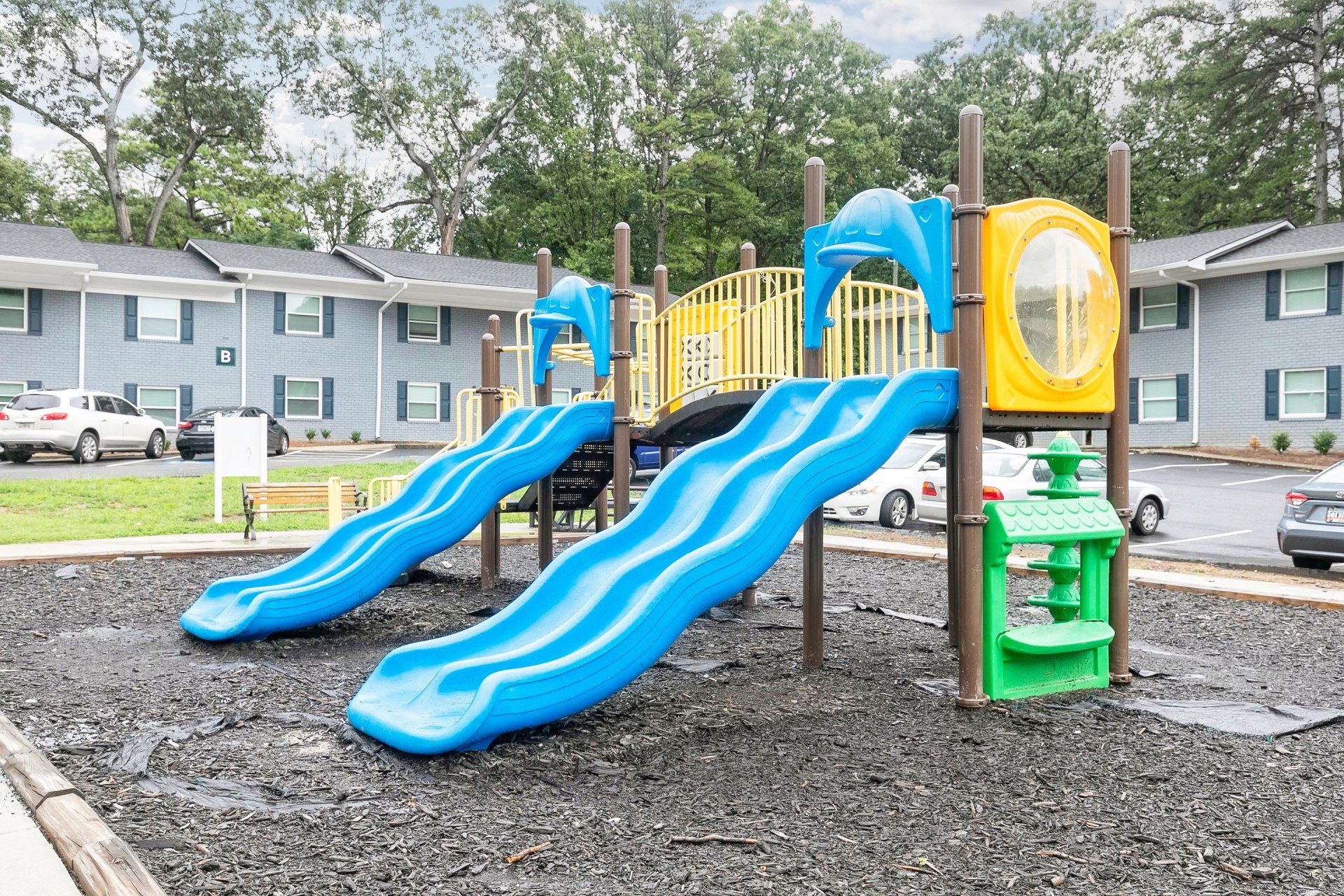 Apartments For Rent In Forest Park, GA - Gardens at Park West - Playground With Slide Set And Bench Near Parking Lot.