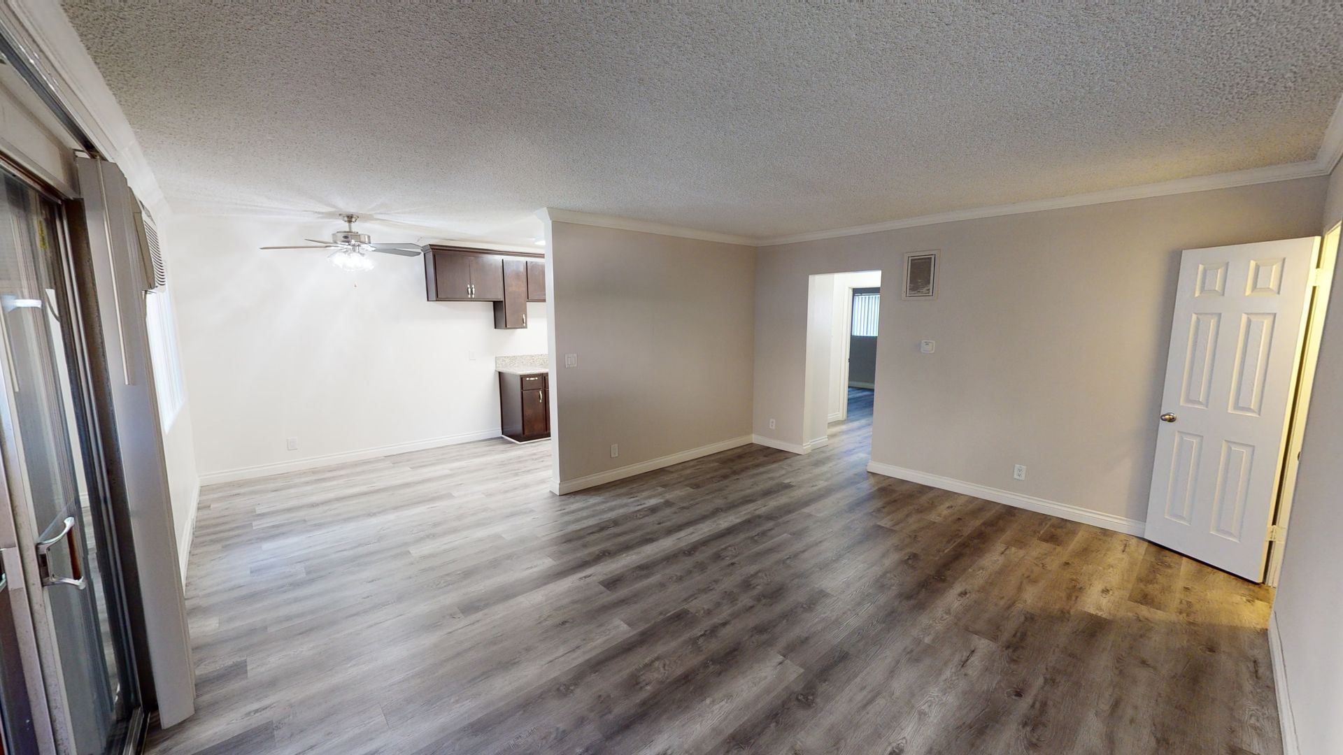 Empty apartment interior with wood-look flooring, neutral walls, and a view into the kitchen.