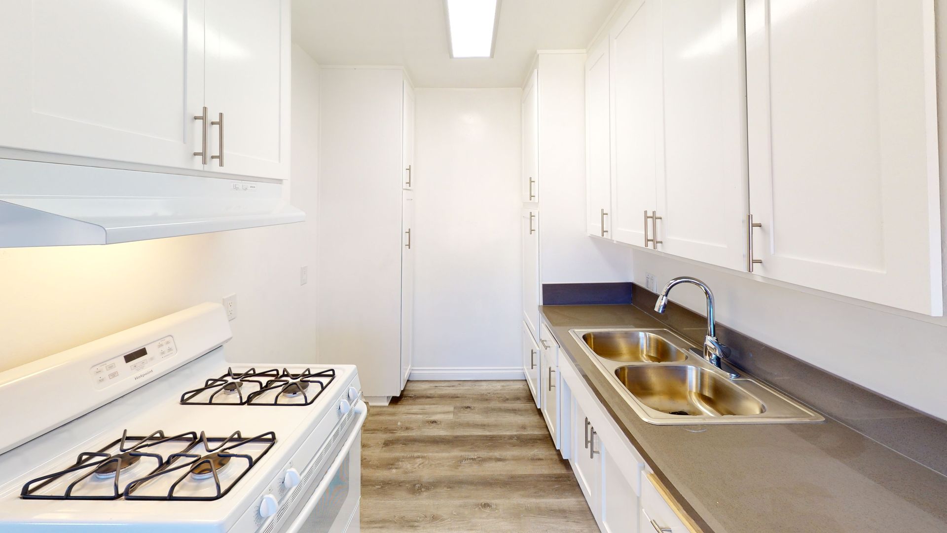 White kitchen with stove, cabinets, sink, and refrigerator. Gray countertops and wood-look flooring.