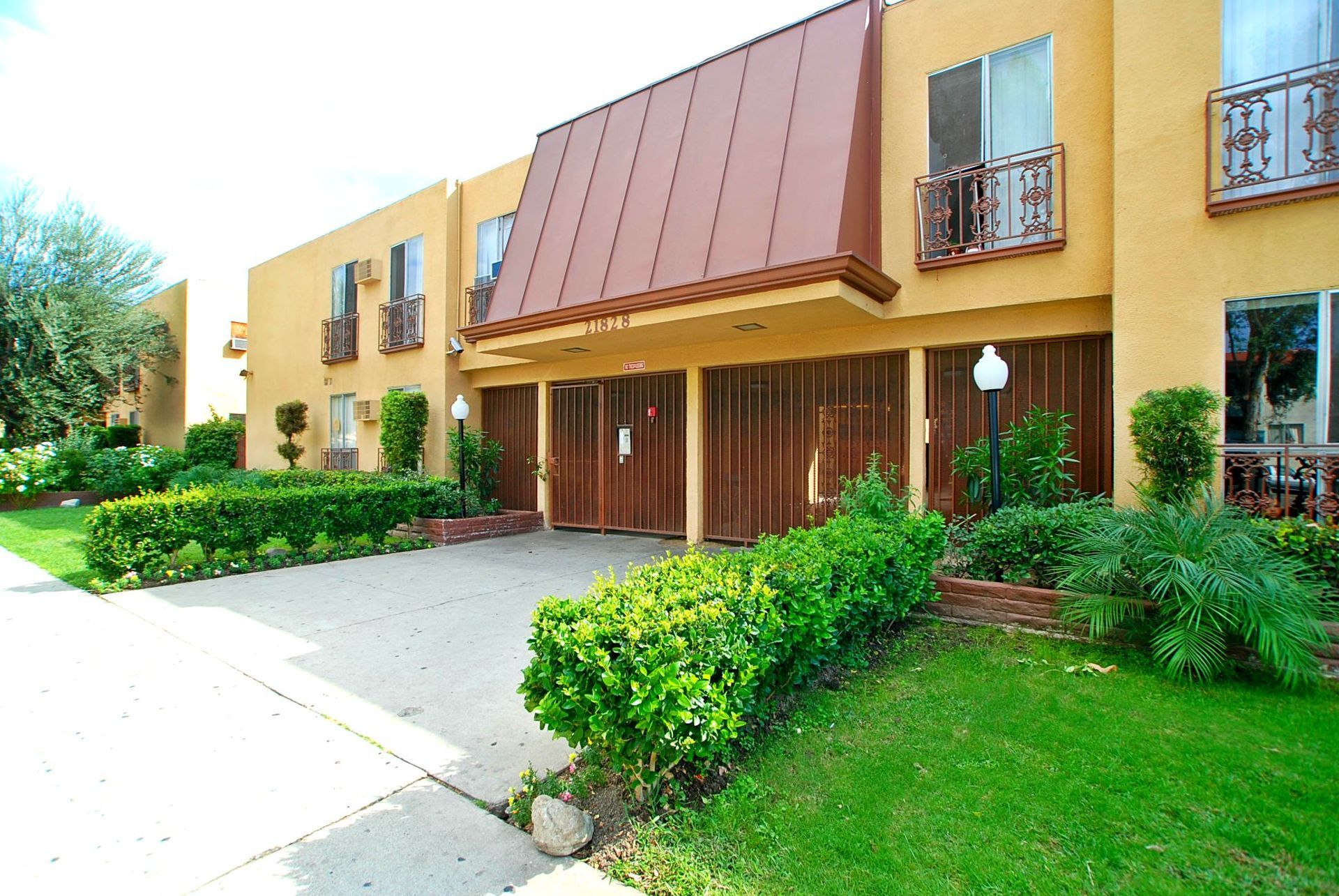 Apartment building with yellow stucco walls, brown garage doors, and a red-brown roof. Lush green lawn and shrubs.