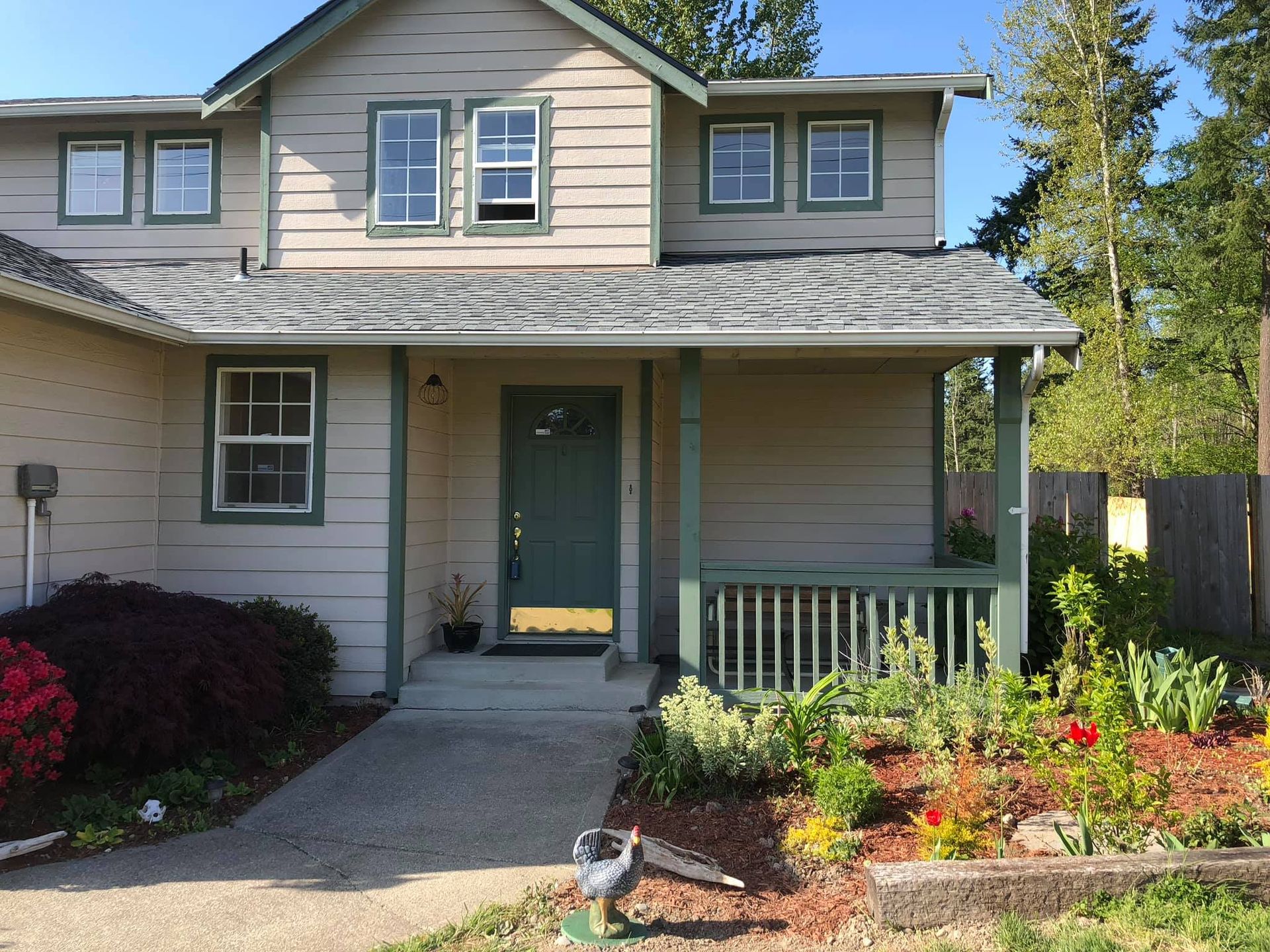 A two-story light-colored house with a dark green front door, a small porch, and a garden in front on a sunny day.