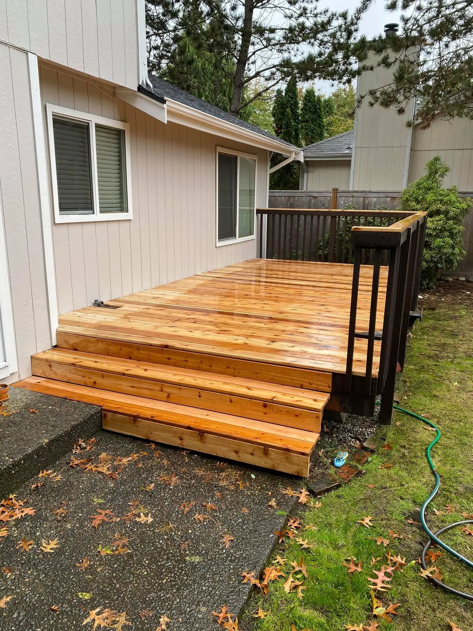 Wooden deck with steps, railing, attached to beige house. Overcast day, wet leaves on concrete.