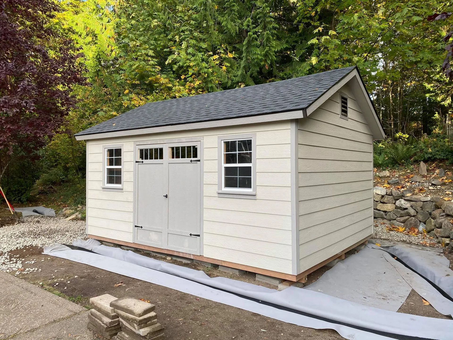 Tan shed with a gray roof and white-trimmed windows and doors, set in a yard with trees.