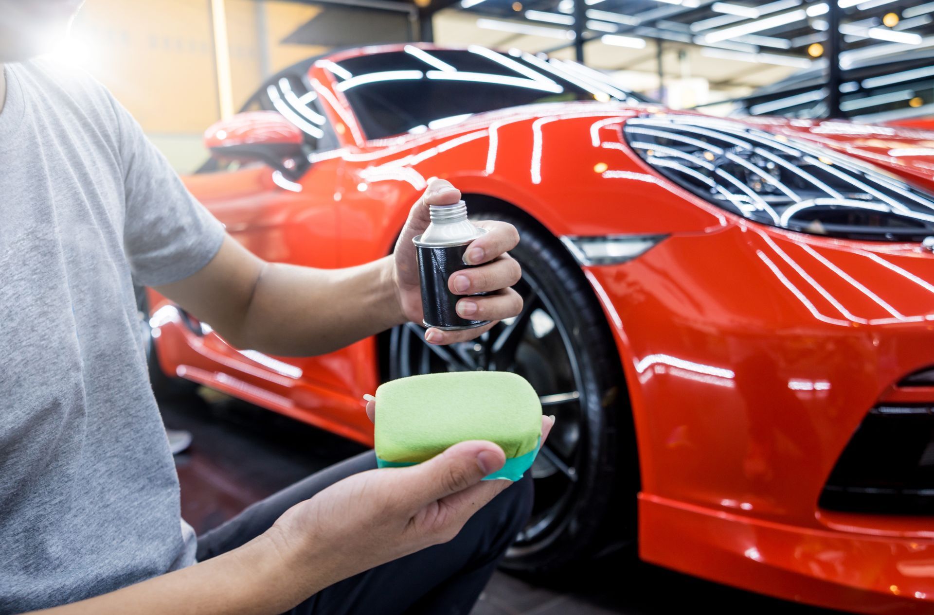 Person spraying a bottle onto a sponge, preparing to detail a shiny red car in a garage.