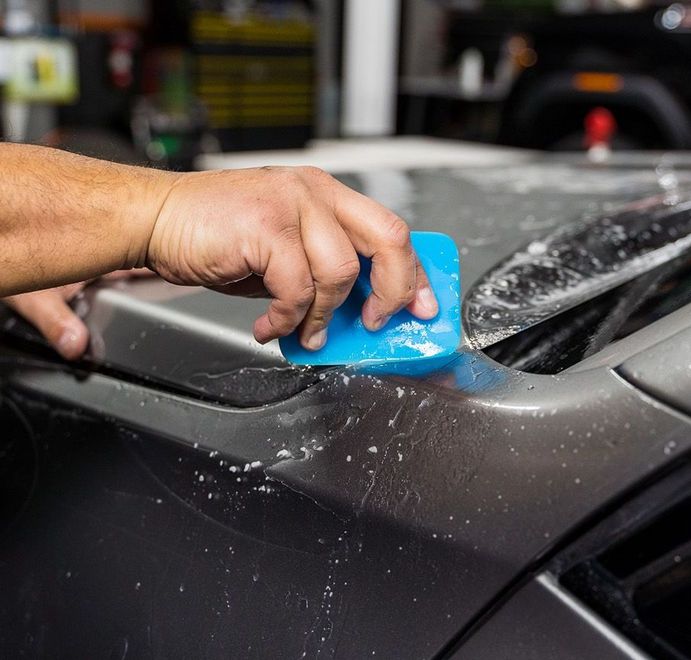 Person applying a protective film to a car's surface with a blue squeegee in a workshop.