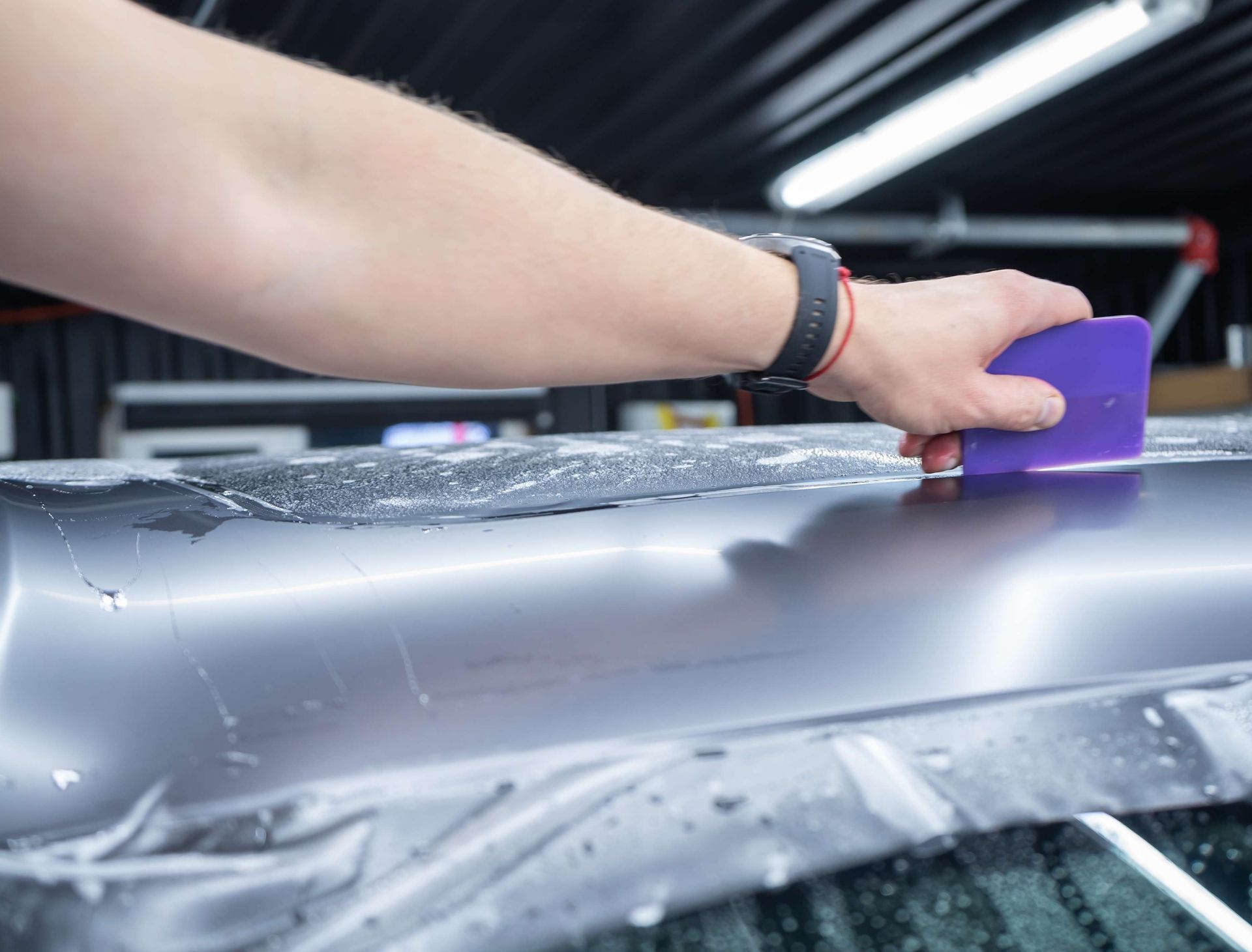 Person smoothing a clear film over a car's surface with a purple squeegee indoors.