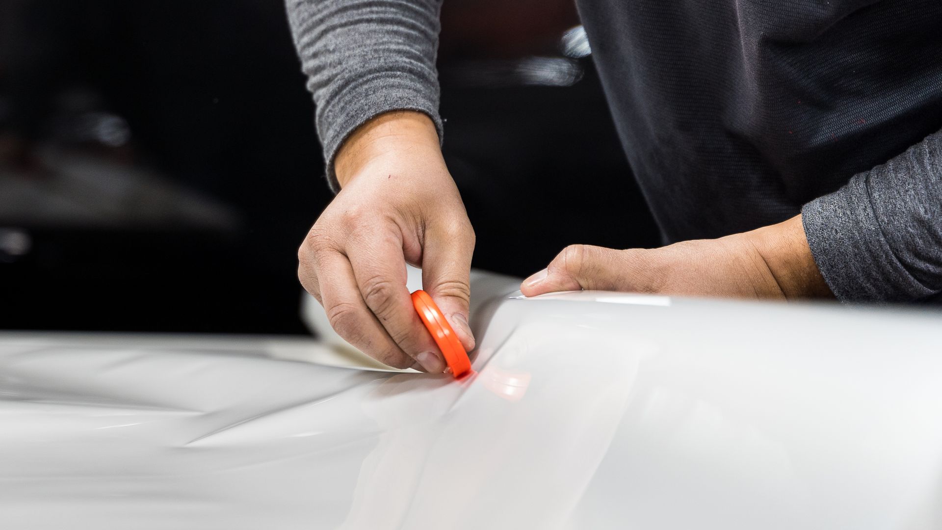 Person using an orange tool on a white car surface, possibly applying film or wrap.