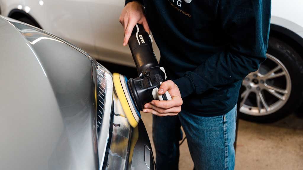Person polishing a silver car fender with a power buffer in a garage.