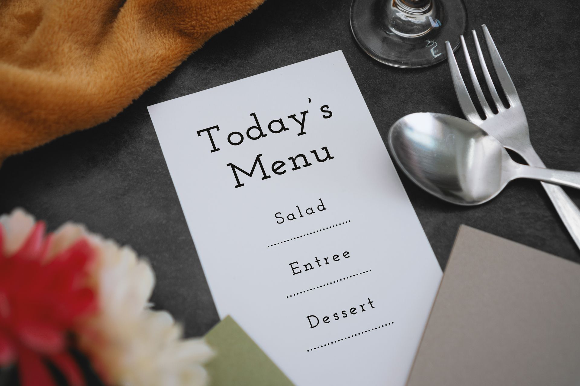 Menu card on a dark surface with silverware, a glass, and flowers nearby.