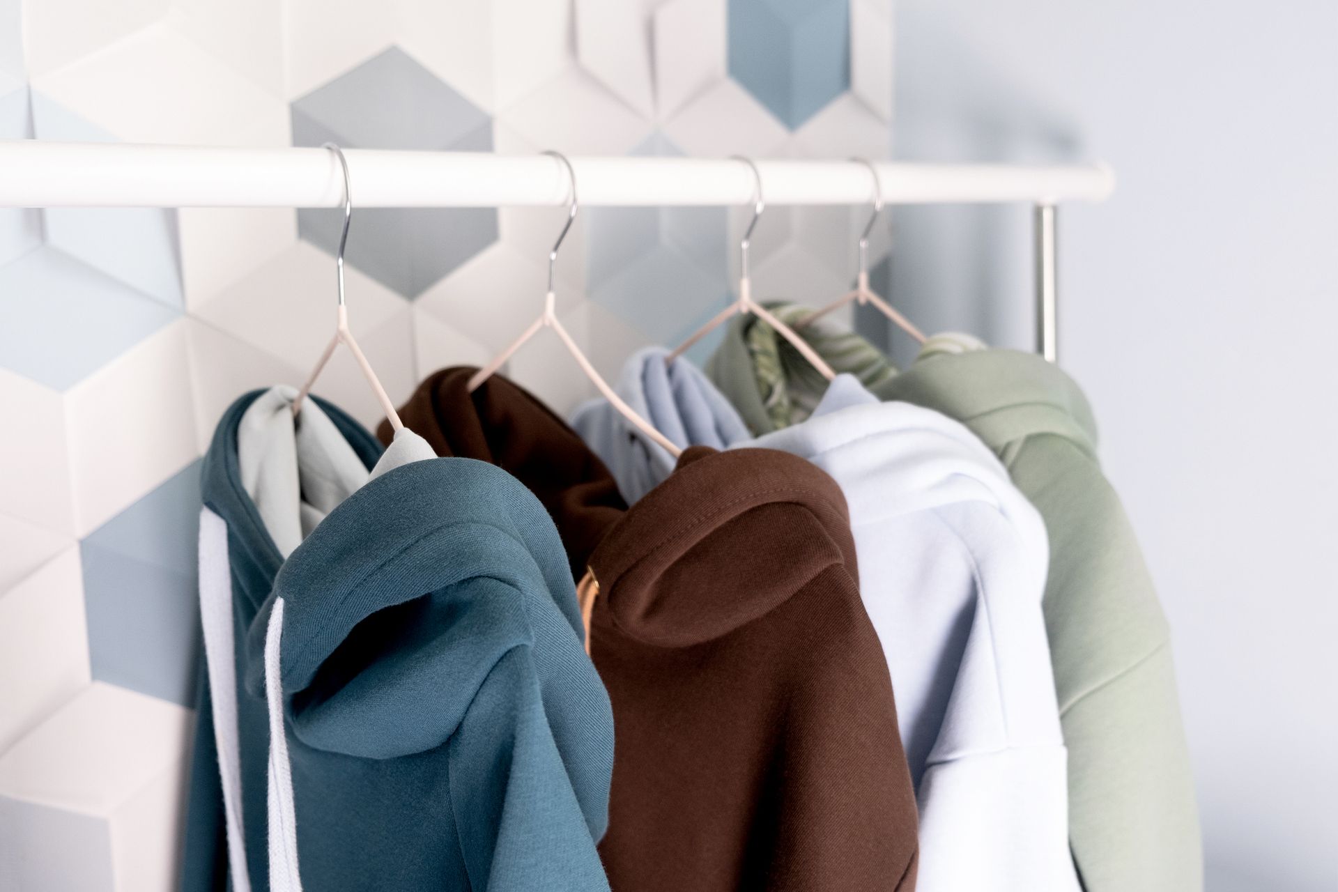 Hoodies in shades of teal, brown, and green hang on a white clothes rack in front of a patterned wall.