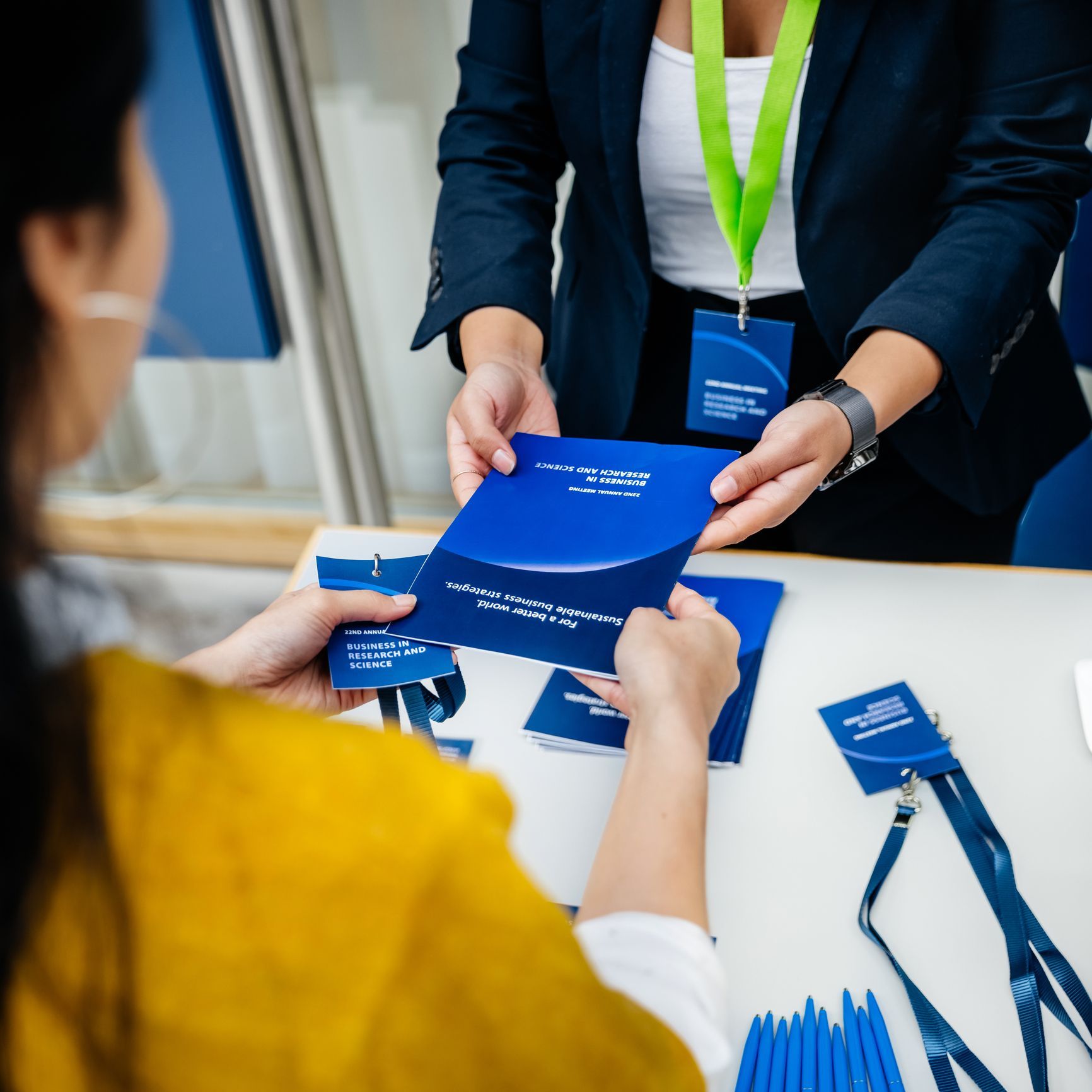 Person in yellow shirt receiving a blue folder from a person in a navy blazer at a registration table.