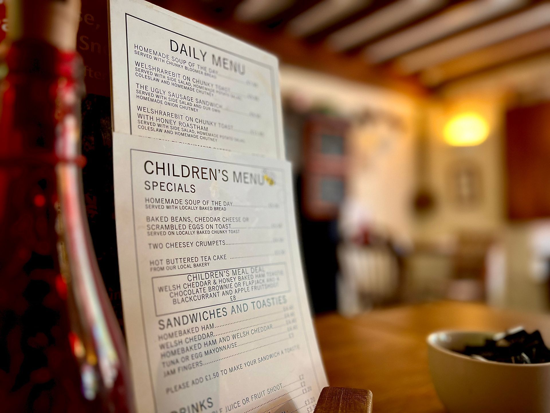 Menu on a wooden table in a cafe, listing daily specials and children's menu. A bowl and bottle are in the foreground.