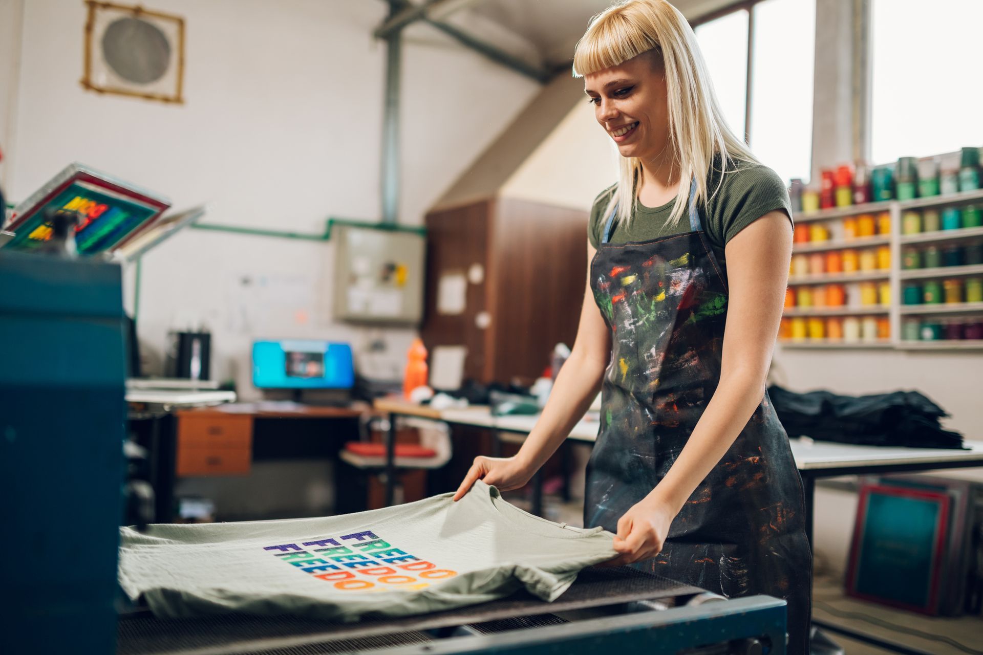 Woman in apron smiles, inspecting a t-shirt freshly printed in a workshop.