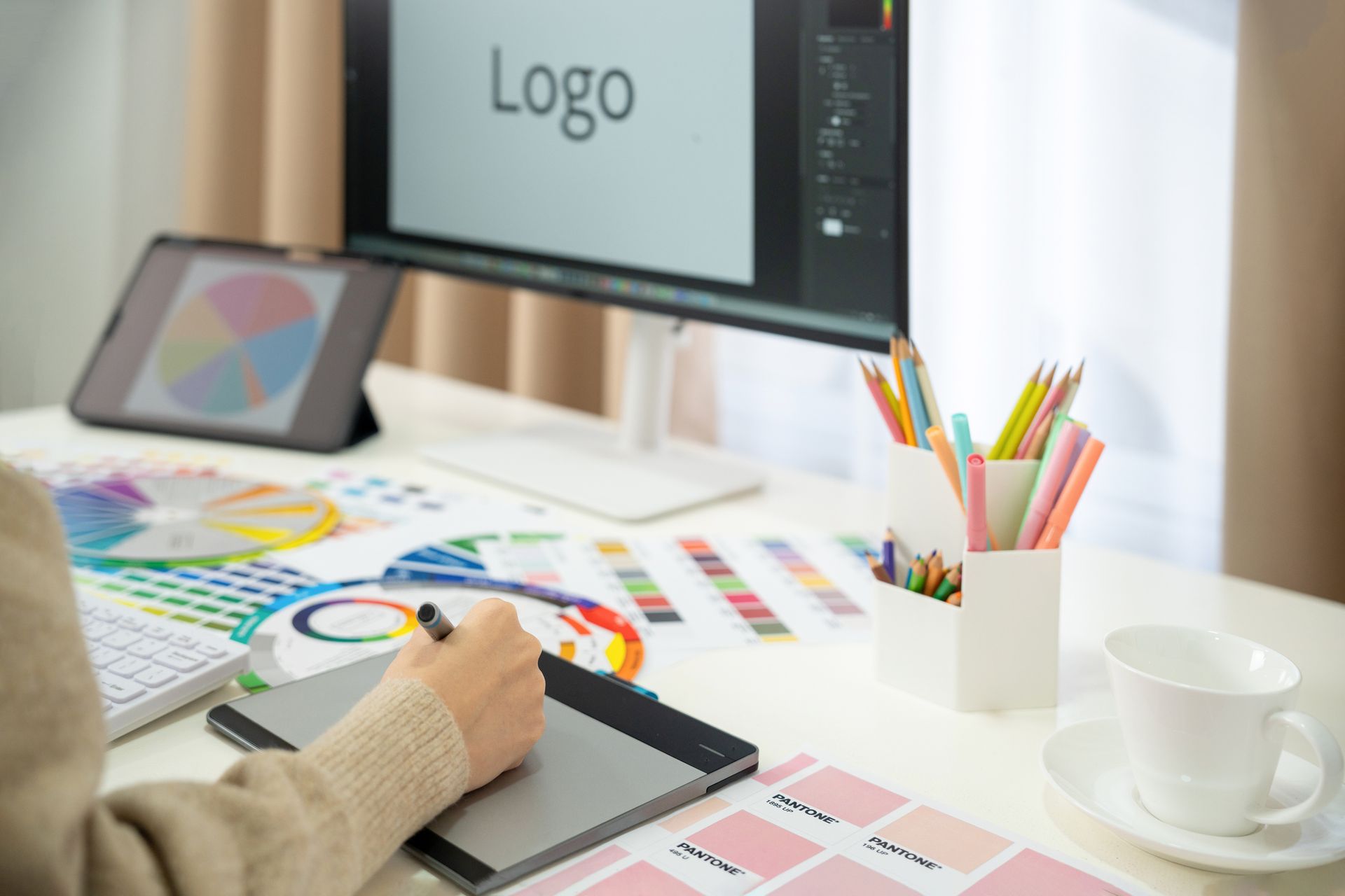 Person using a graphics tablet and computer to design a logo, surrounded by color palettes and art supplies.