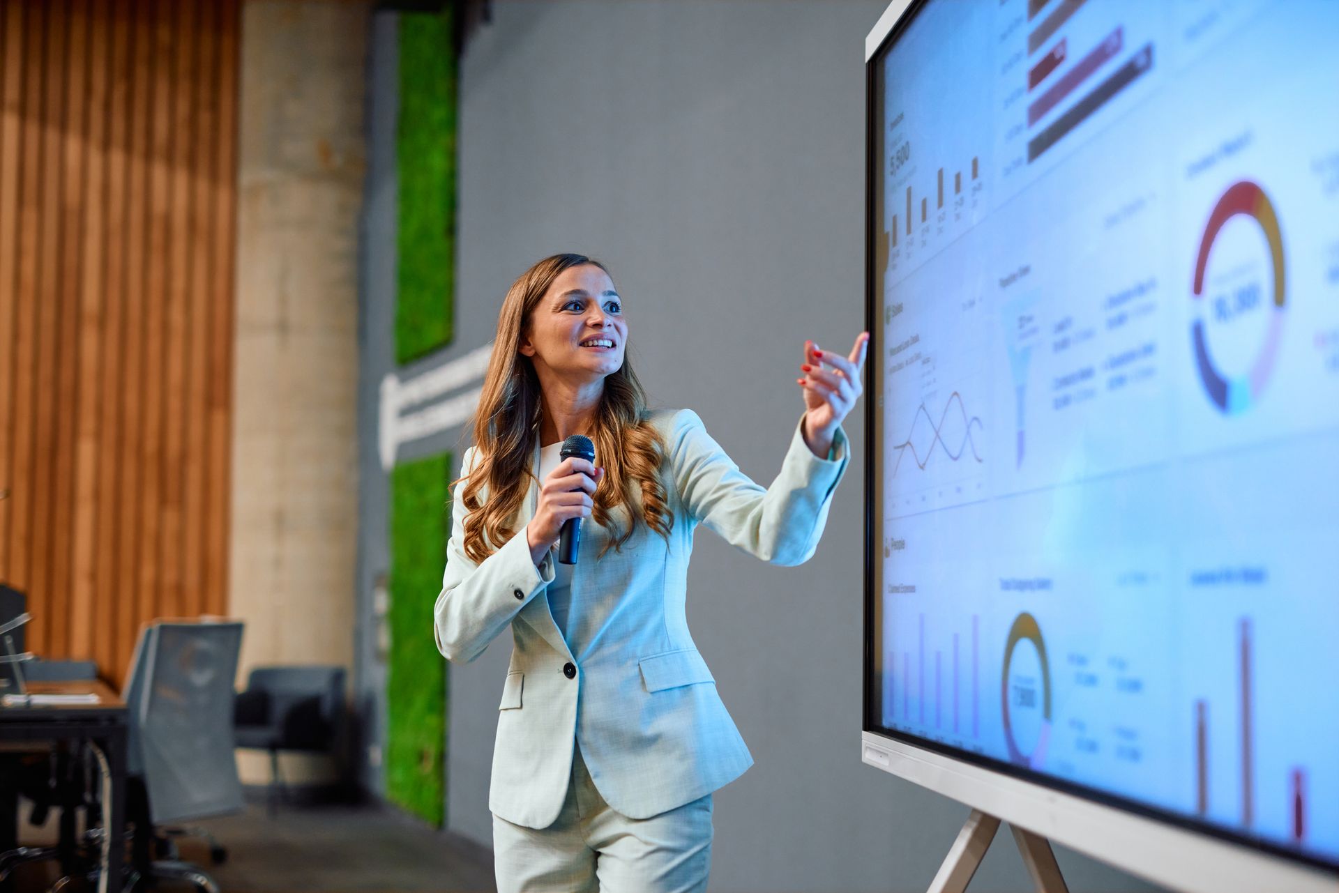 Woman presenting data on a screen, holding a microphone. Light blue suit in an office setting.