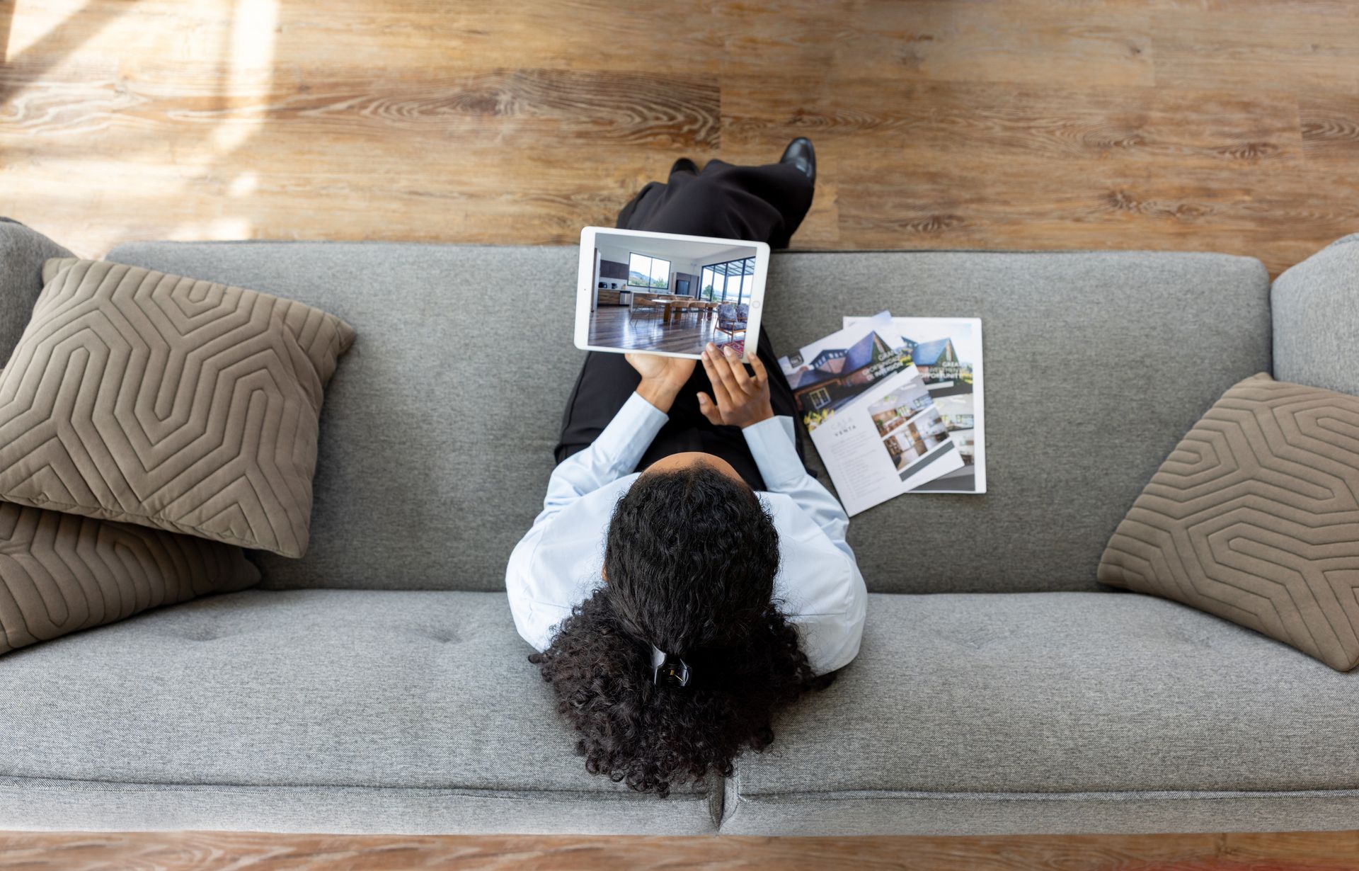 Woman seated on gray couch, using tablet and looking at magazine. Wooden floor in background.
