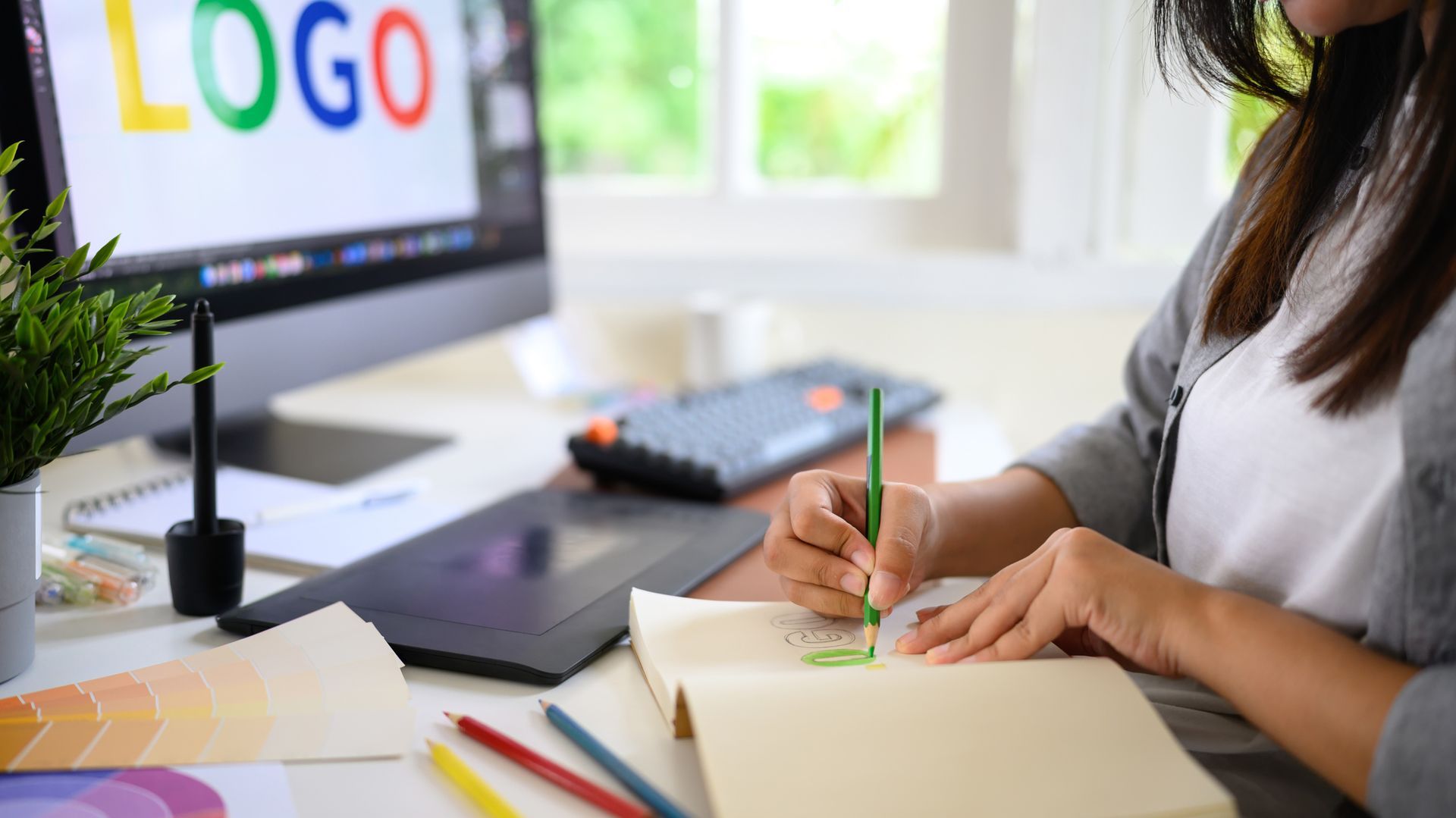 Person sketching a design with a green pencil, next to a computer displaying a colorful logo.