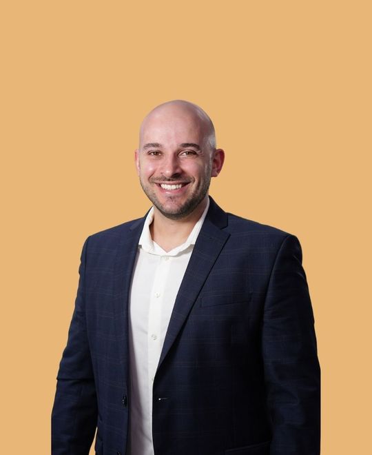 Smiling bald man in a navy blazer and white shirt against an orange background.