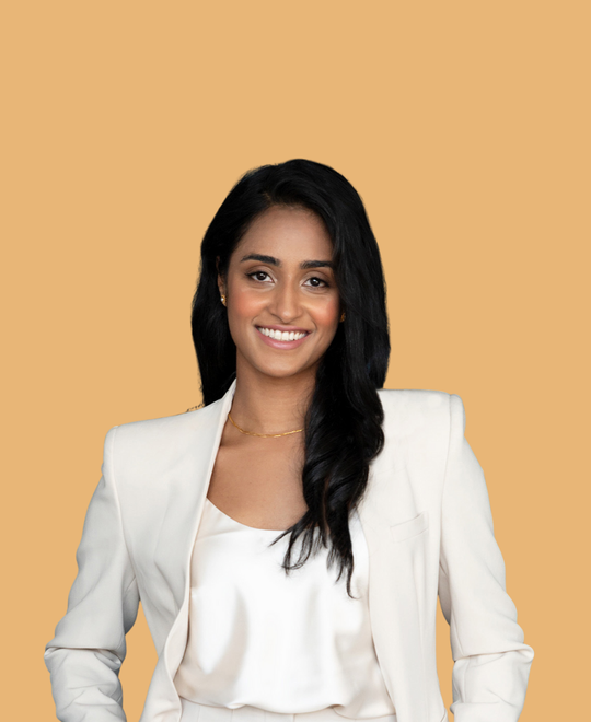 Woman with dark hair smiles, wearing a white blazer and top against a peach background.