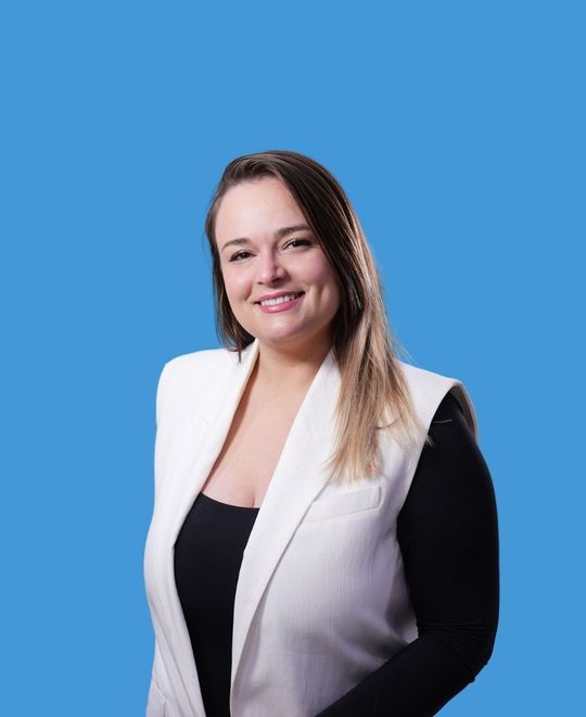 Woman with long brown hair smiles, wearing a white vest over a black top, against a blue background.