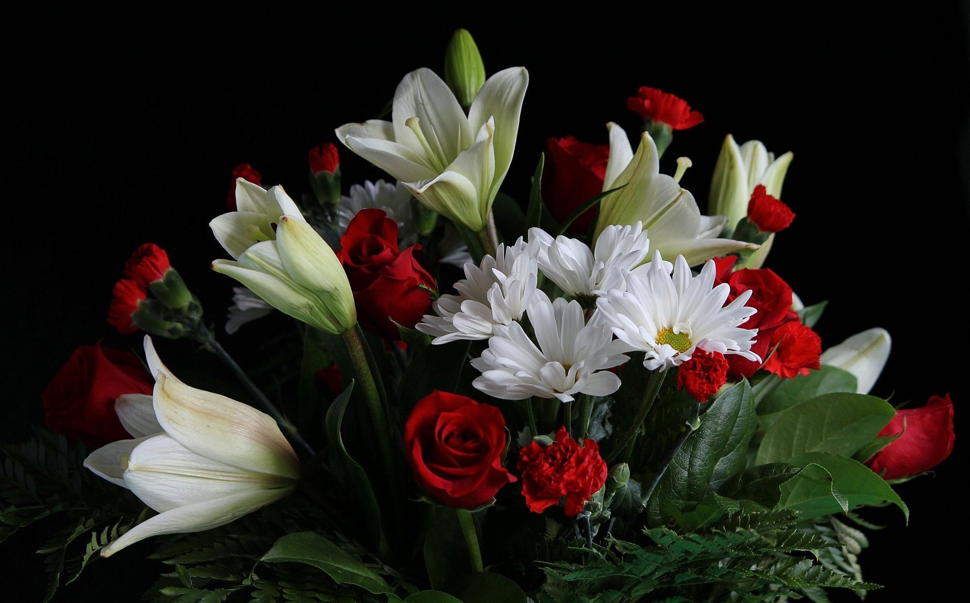 Bouquet of white lilies, red roses, carnations, and white daisies against a black background.