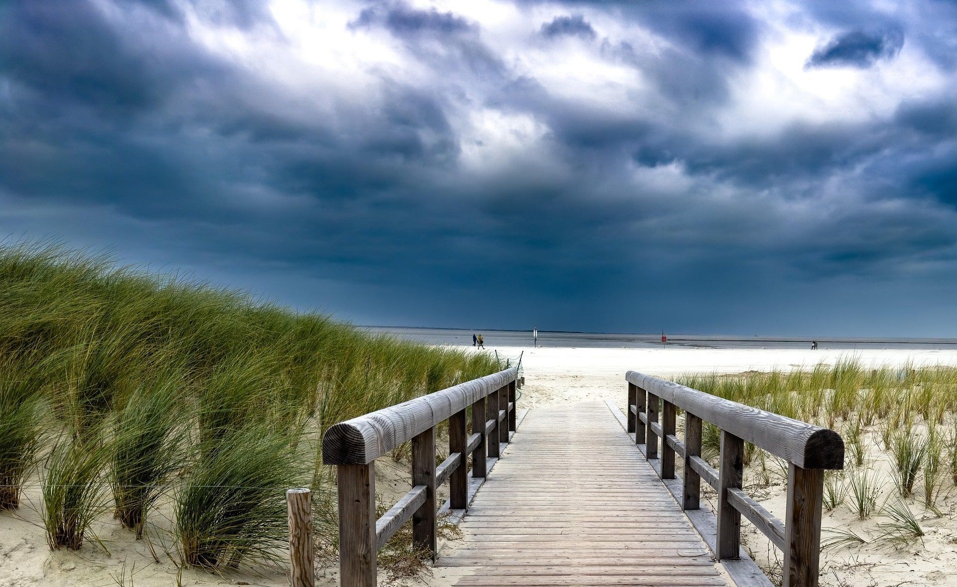 Wooden boardwalk on a sandy beach, flanked by green grass, under a dark, cloudy sky.