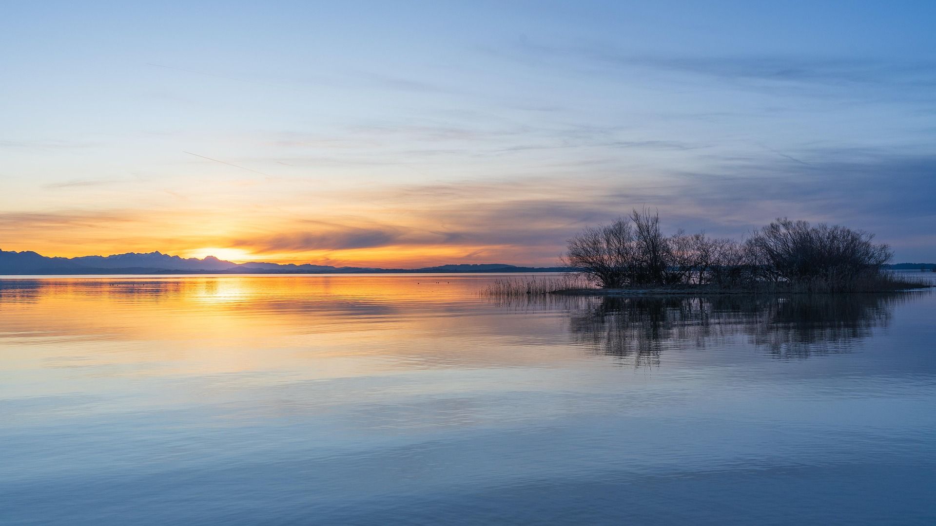Sunset over a calm lake with a small island and reflective colors of orange, yellow, and blue.