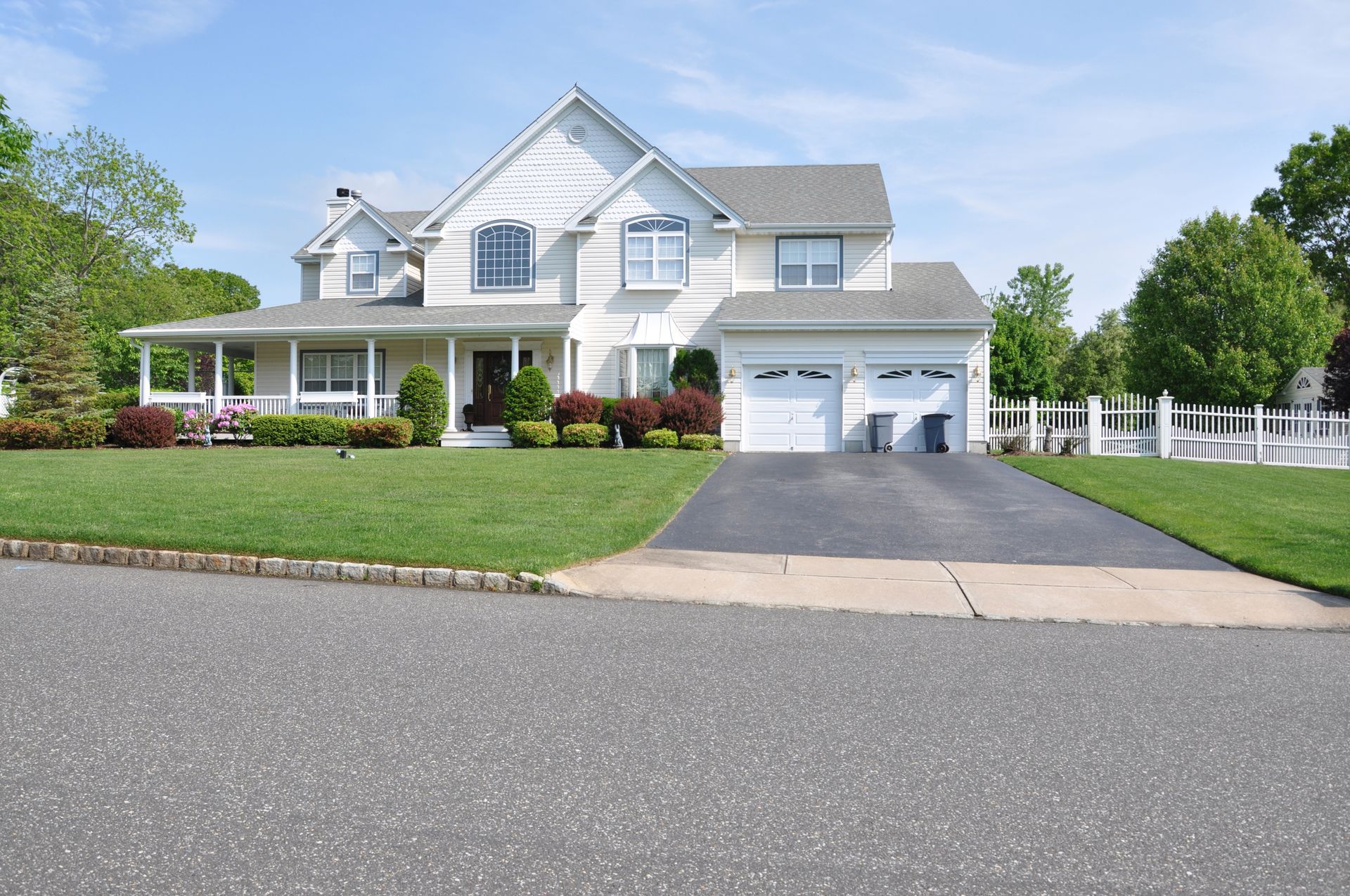 White two-story house with a green lawn, two-car garage, and black asphalt driveway on a sunny day.
