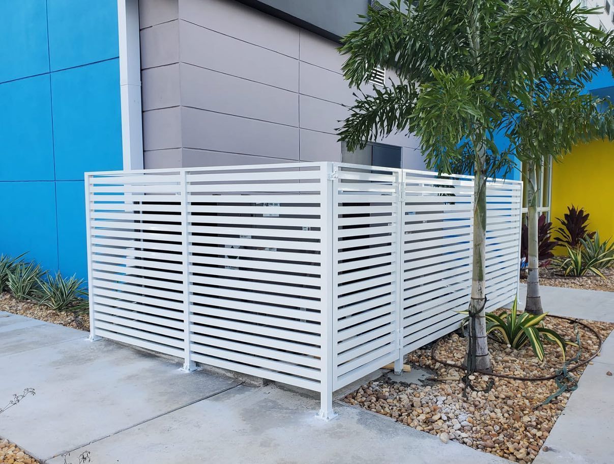 A white fence is surrounding an air conditioner outside of a building.