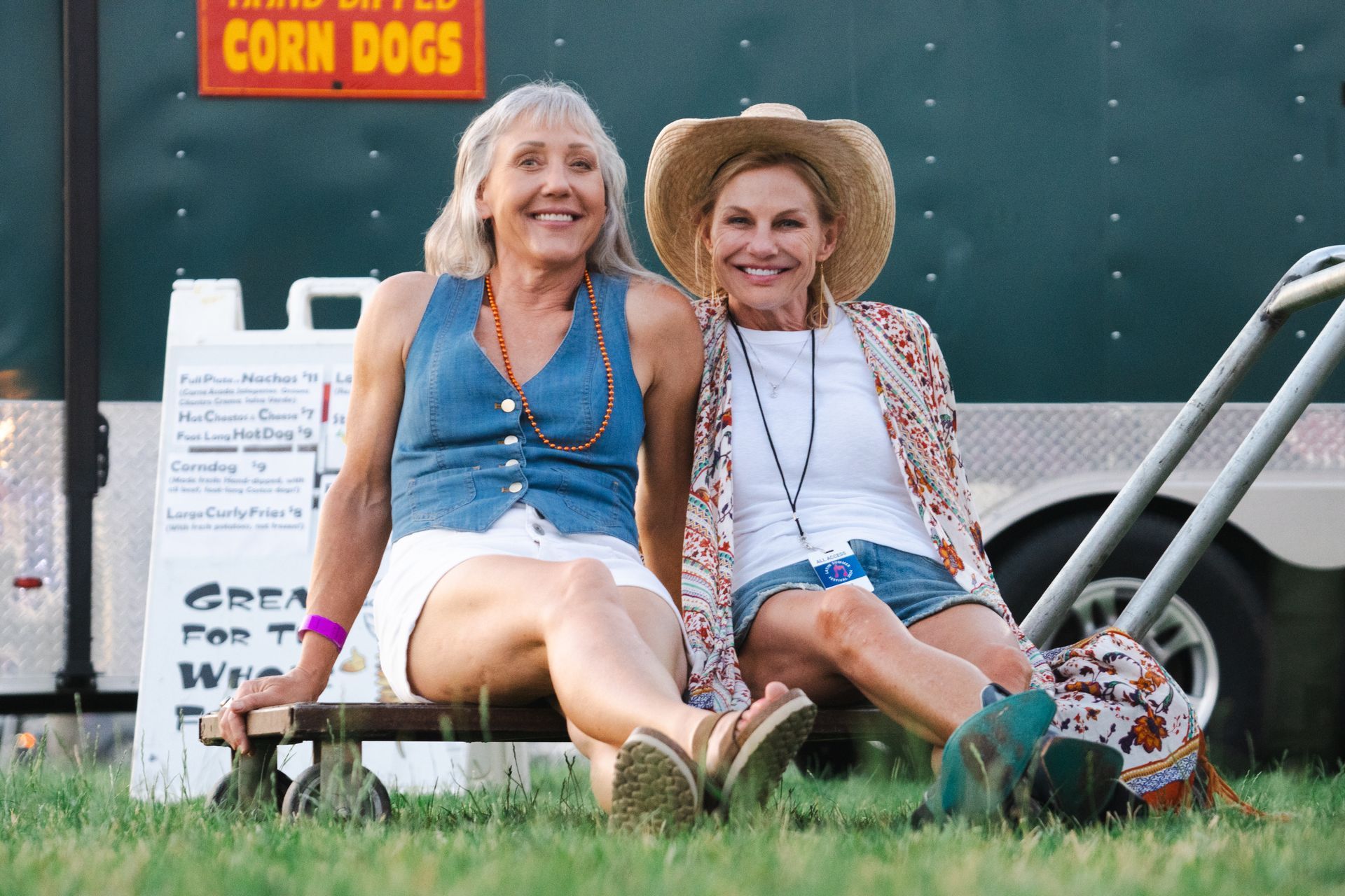 Two women are sitting next to each other in front of a truck that says corn dogs.