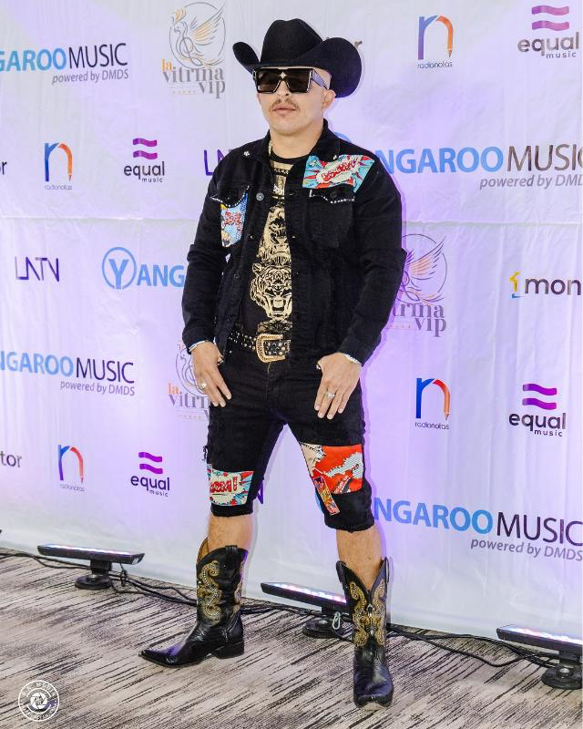 A man wearing a cowboy hat and cowboy boots is standing on a red carpet.