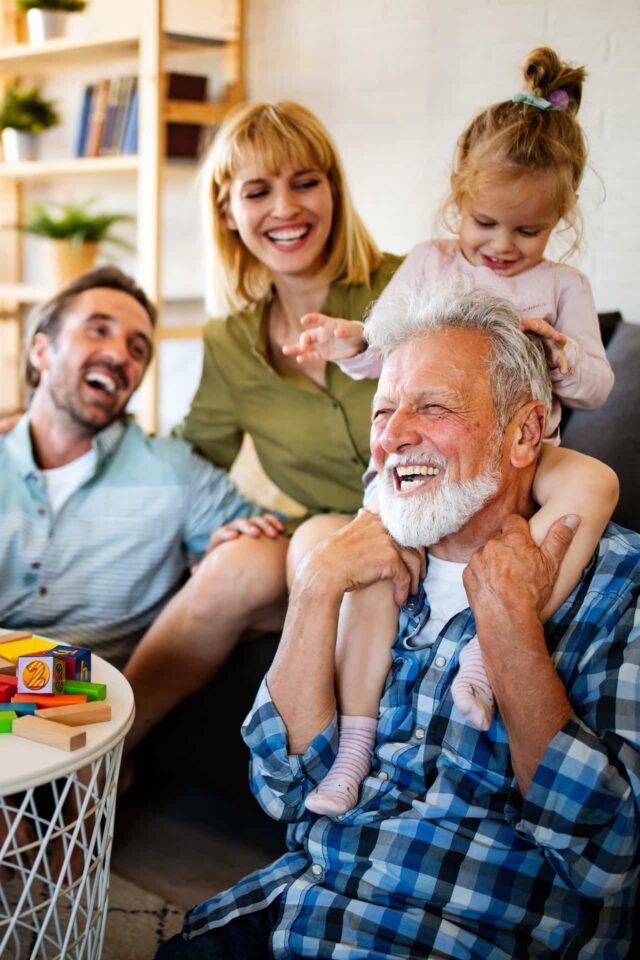 Family smiling together on a couch, with a toddler on an older man's shoulders in a cozy living room.
