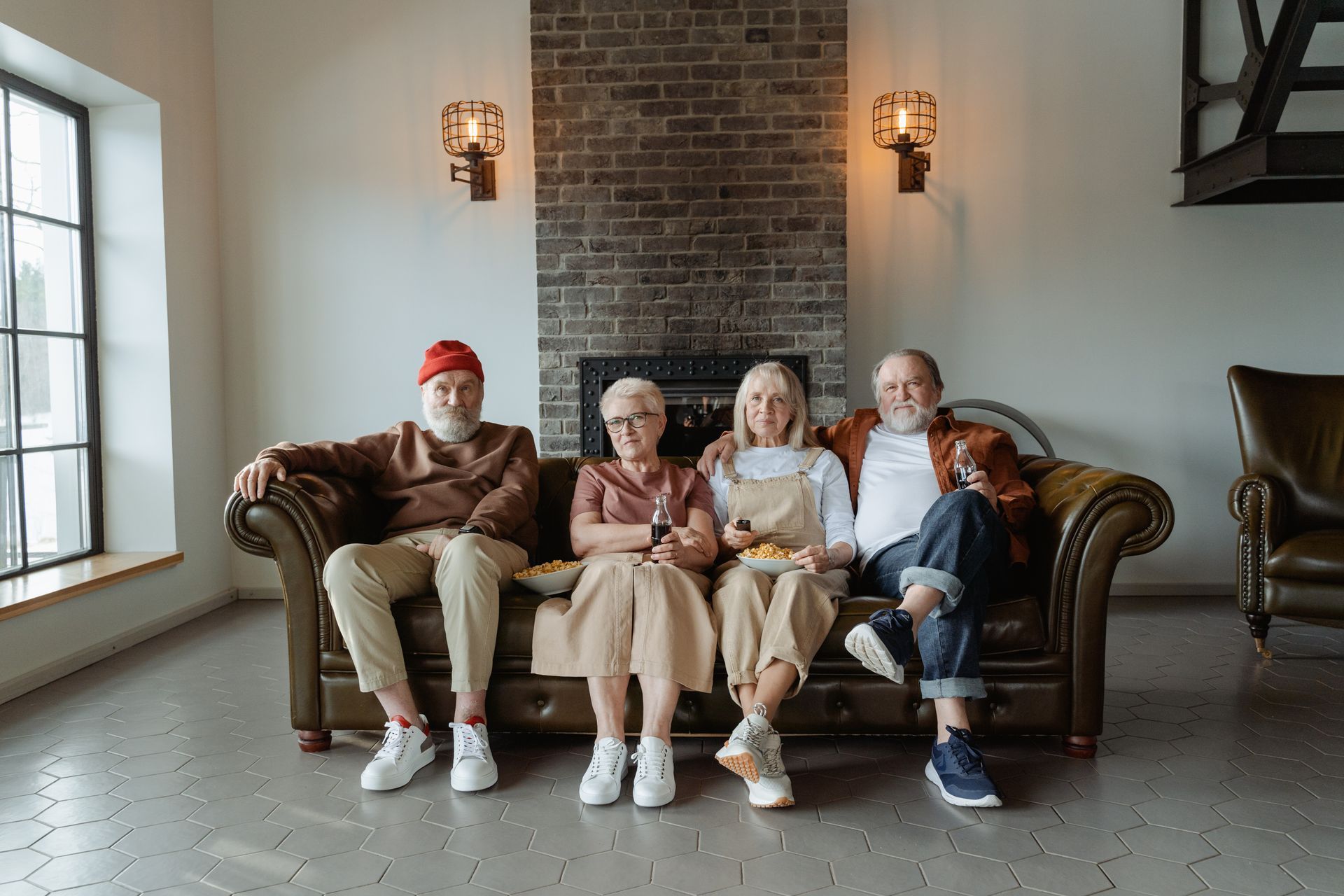 Four people sitting on a brown couch in a bright living room, with wall sconces and a window nearby.