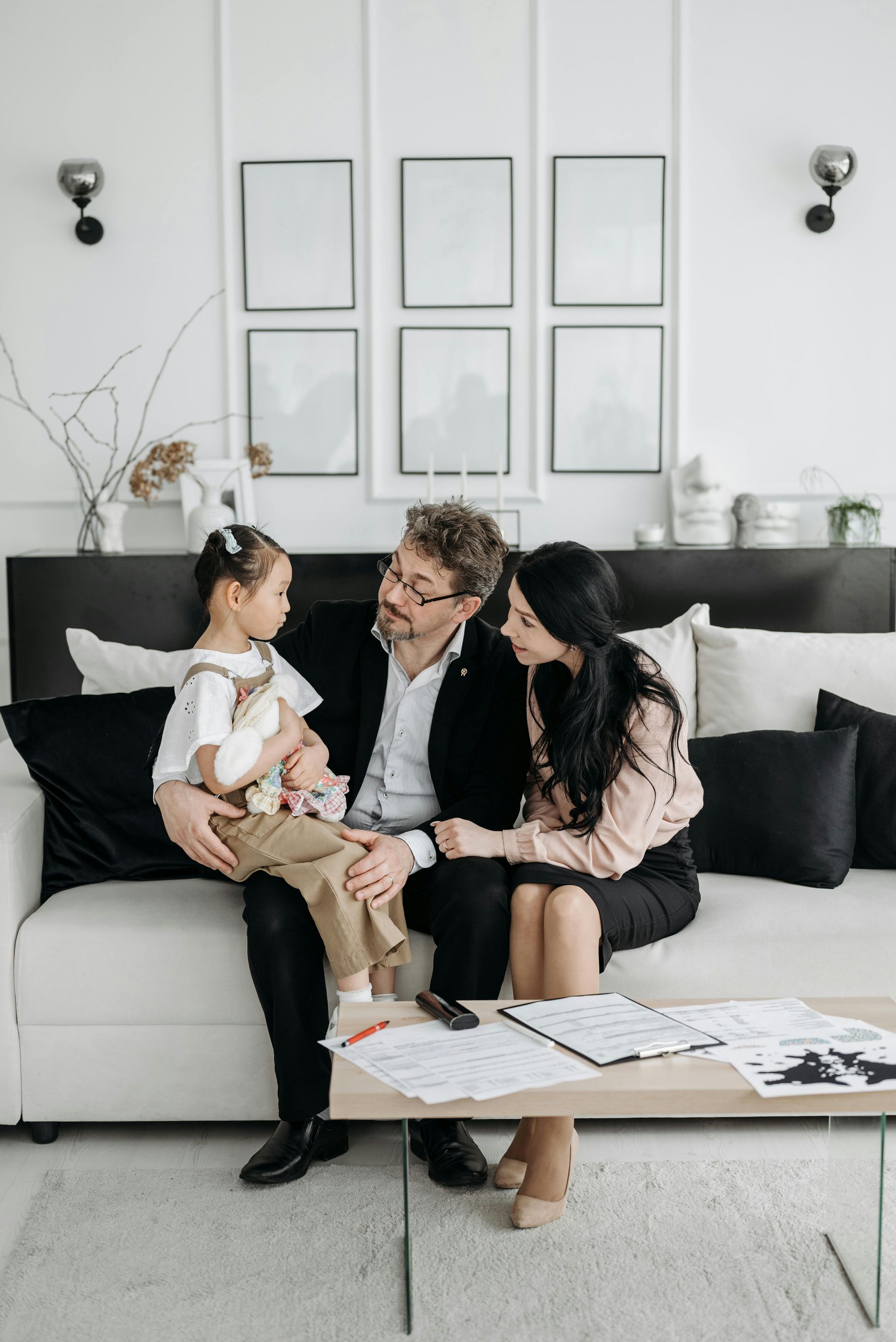 Three people sitting on a couch in a bright living room, looking at a baby on the left.
