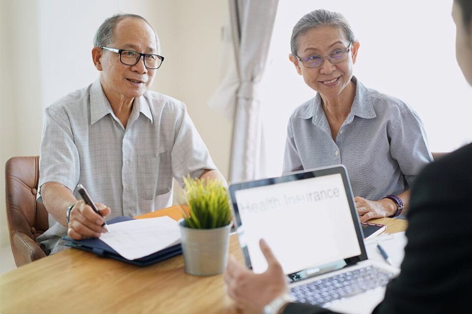 Two people meeting at a desk with a laptop, notebook, and pen in a bright office.