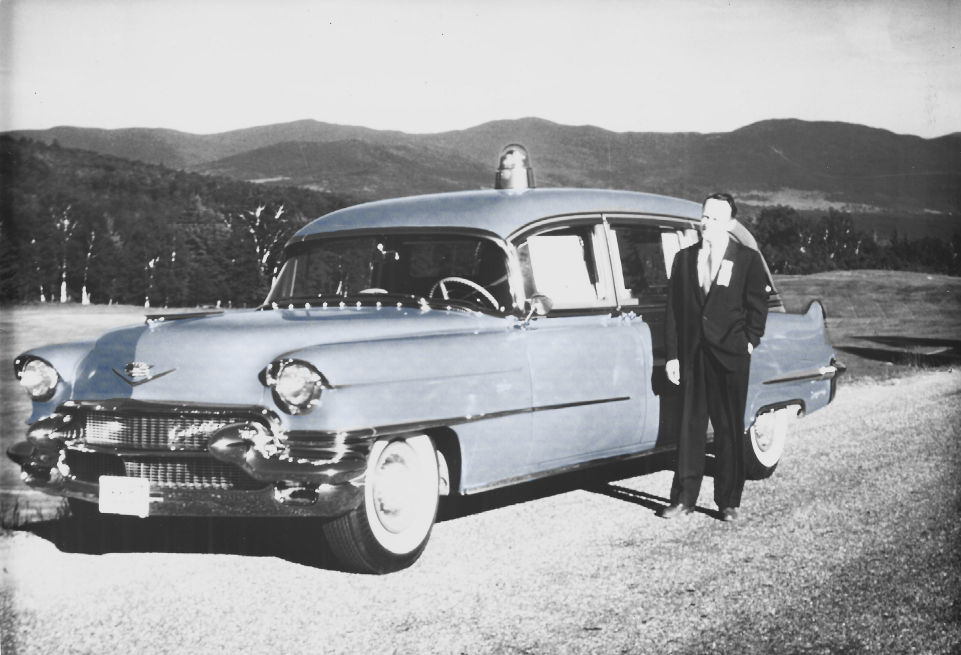 Man standing by a light blue 1950s Cadillac ambulance, mountains in the background.