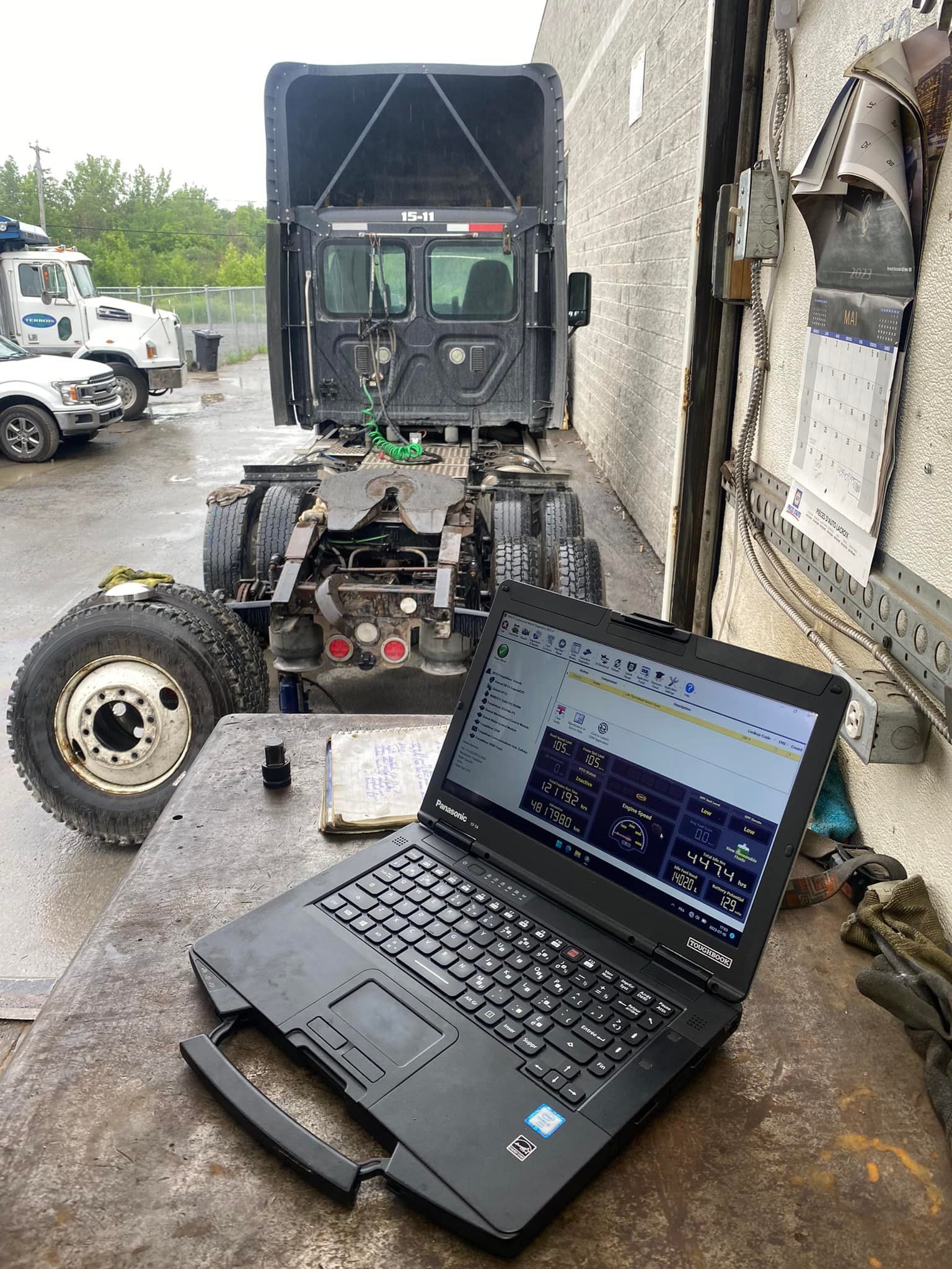 Laptop displaying diagnostics sits on a table, facing a truck chassis with a partially dismantled cab.