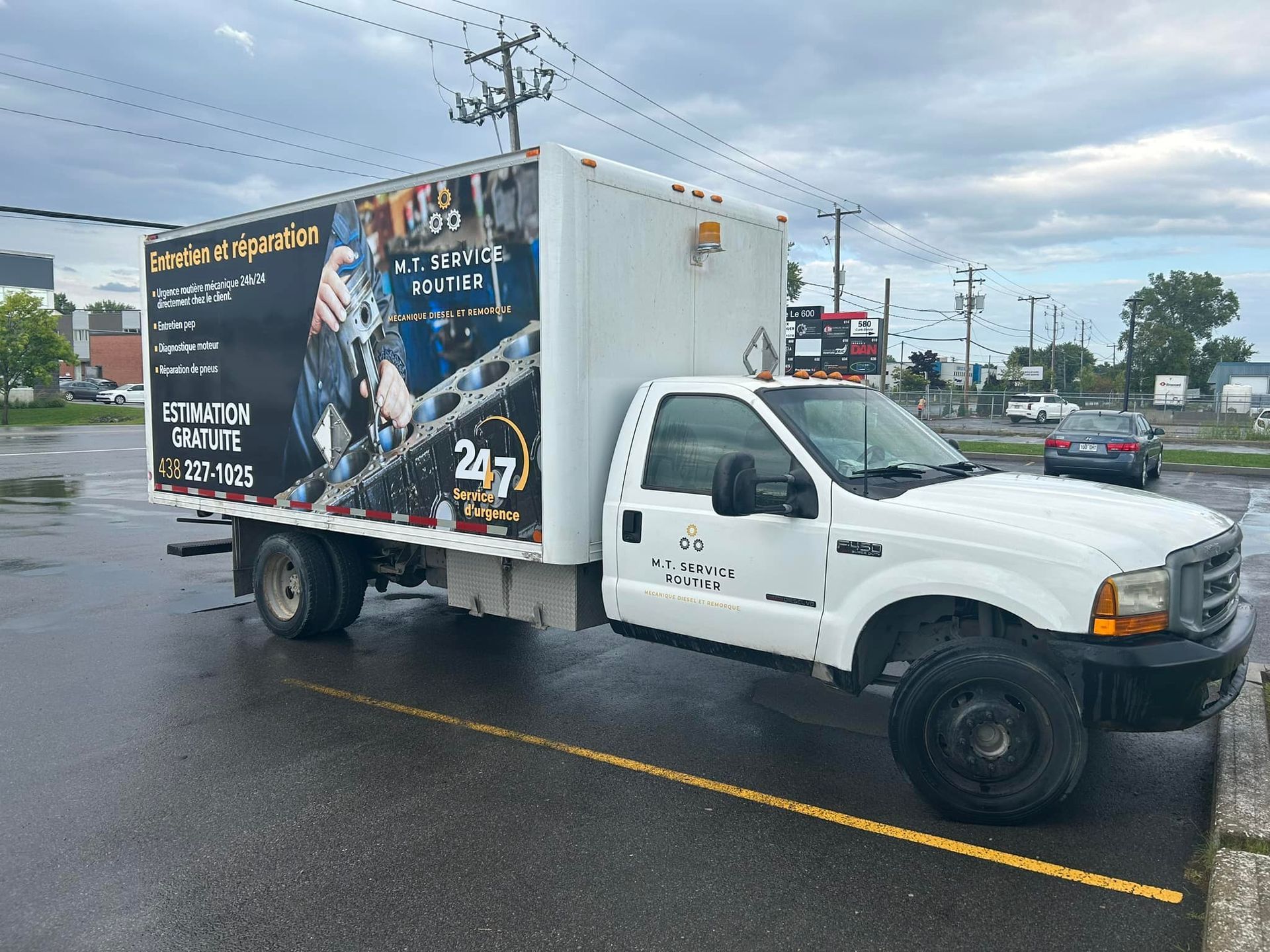 Camionnette blanche avec une grande caisse à l'arrière, garée sur un parking asphalté mouillé sous un ciel nuageux.
