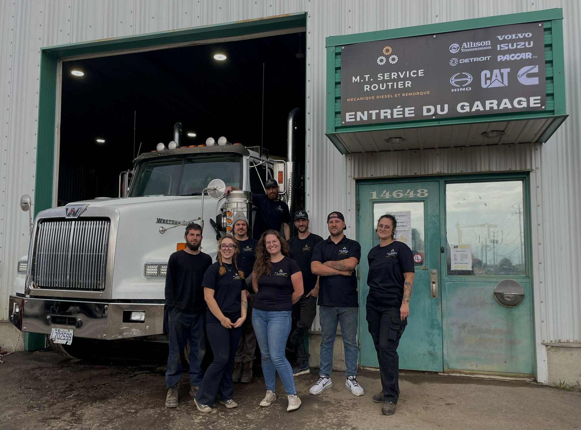 Un groupe de mécaniciens devant un garage et un gros camion blanc. Ils portent des uniformes de travail et sourient.