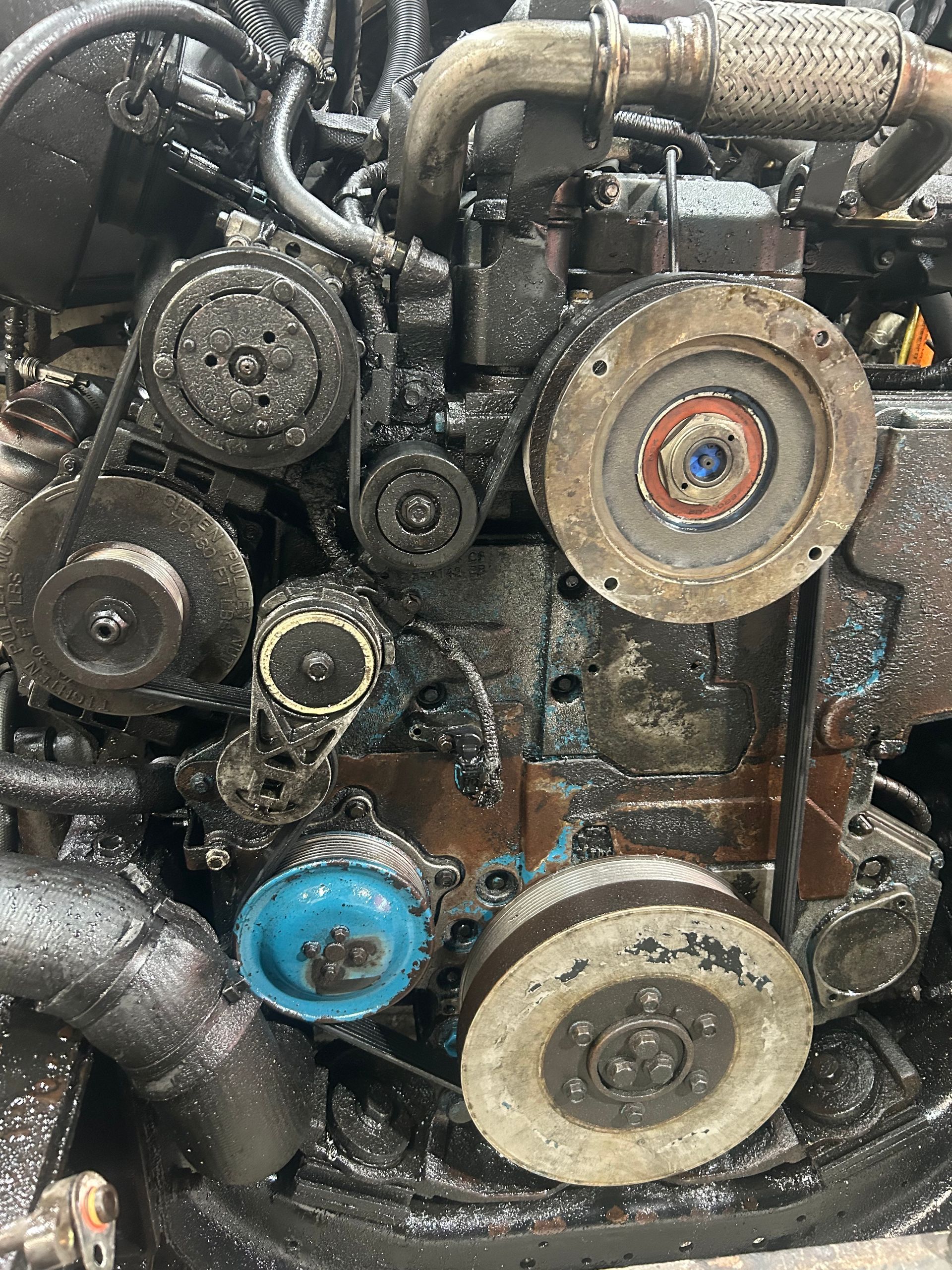 Moteur noir et huileux avec plusieurs poulies, courroies et tuyaux ; semble être dans un cadre mécanique.