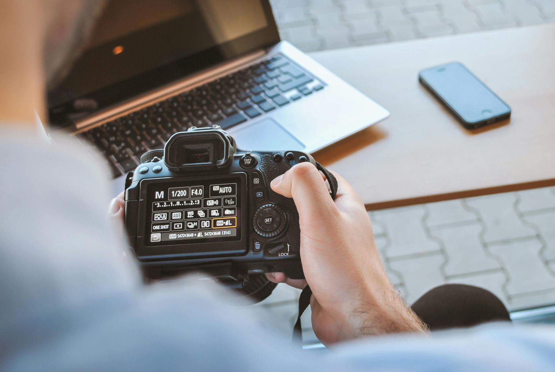 Une personne tient un appareil photo et regarde l'écran. Ordinateur portable et téléphone posés sur une table en arrière-plan.