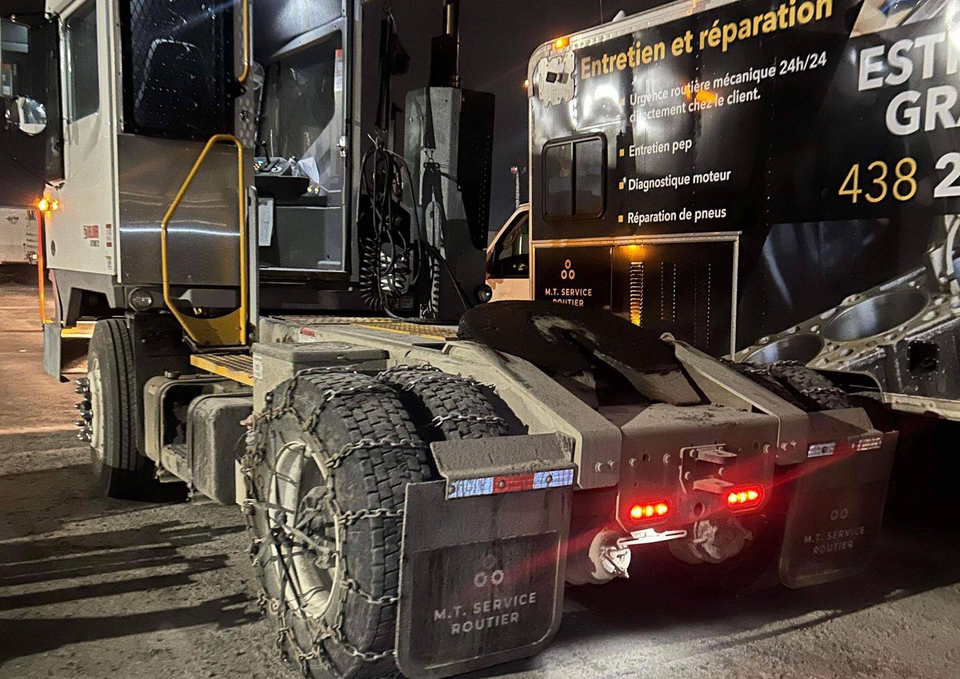 Camion lourd avec chaînes à neige, stationné la nuit. Détails : métal argenté, feux rouges et atelier de réparation.