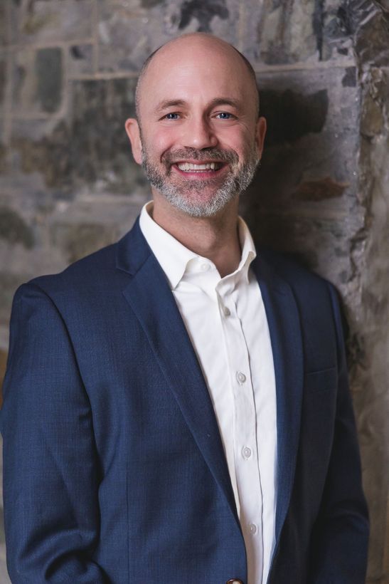 Jed looking at the camera, smiling, in a blue blazer and white collared shirt. He is standing in front of a stone wall.