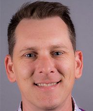 A man is smiling for the camera and wearing a purple shirt.