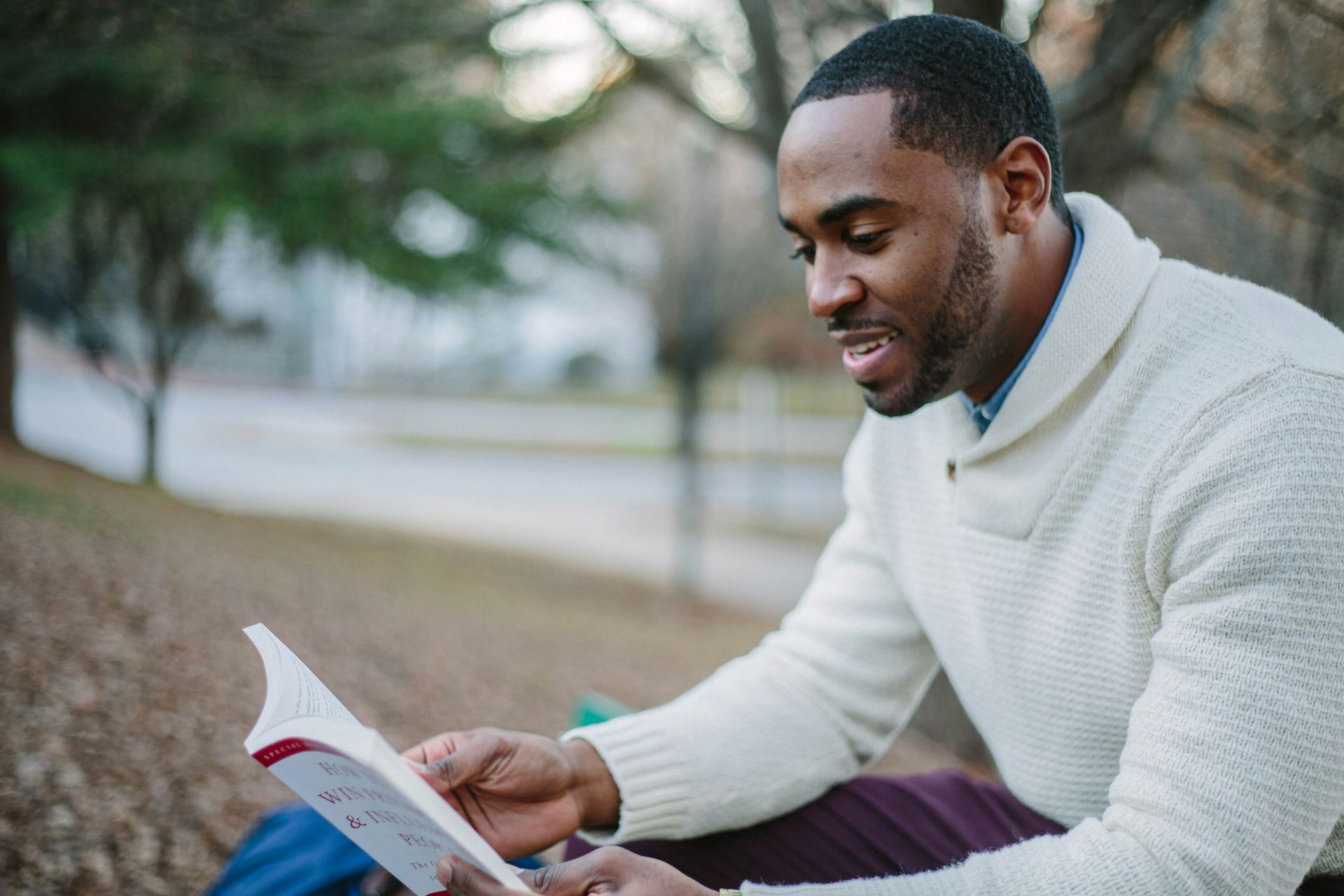 A man in a white sweater is sitting under a tree reading a book.