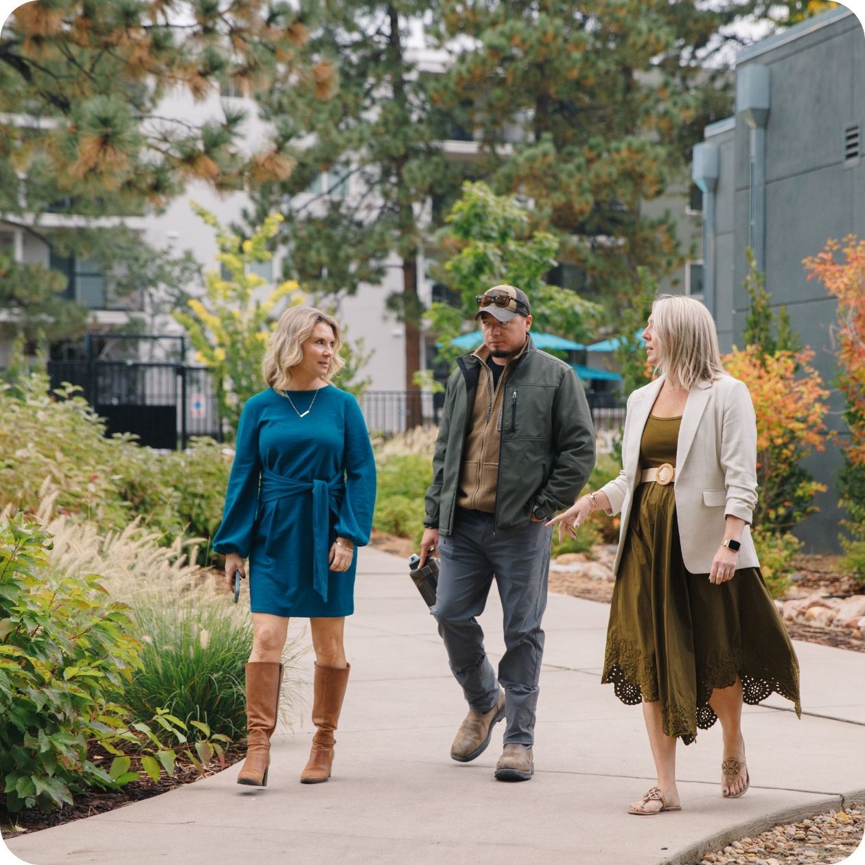 Three people walking on a path outdoors, smiling. Green trees, building in background.