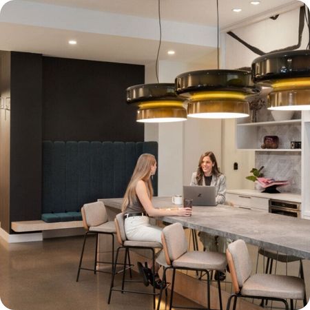 Two people working at a long table in a modern office, under large pendant lights.