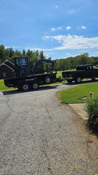 Black truck pulling a trailer with heavy machinery on a gravel driveway, against a sunny, blue sky.